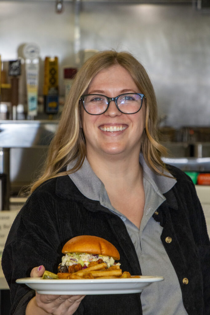Caitlin is holding a burger while looking at the camera and smiling. She is wearing a grey polo shirt with a black jacket. She is wearing black rimmed glasses.